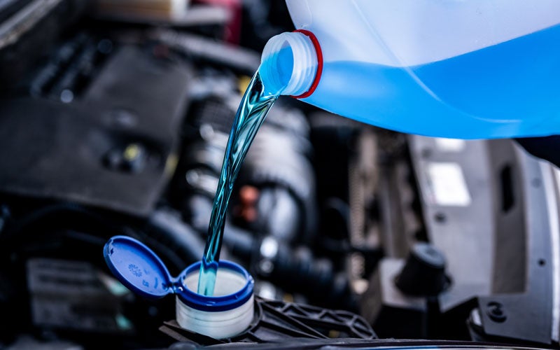 Blue windshield washer fluid being poured from a plastic container into a vehicle’s washer